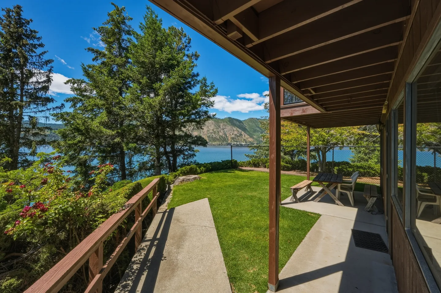 A covered concrete patio with a picnic table looking out onto a grassy yard with views of a lake and mountains.