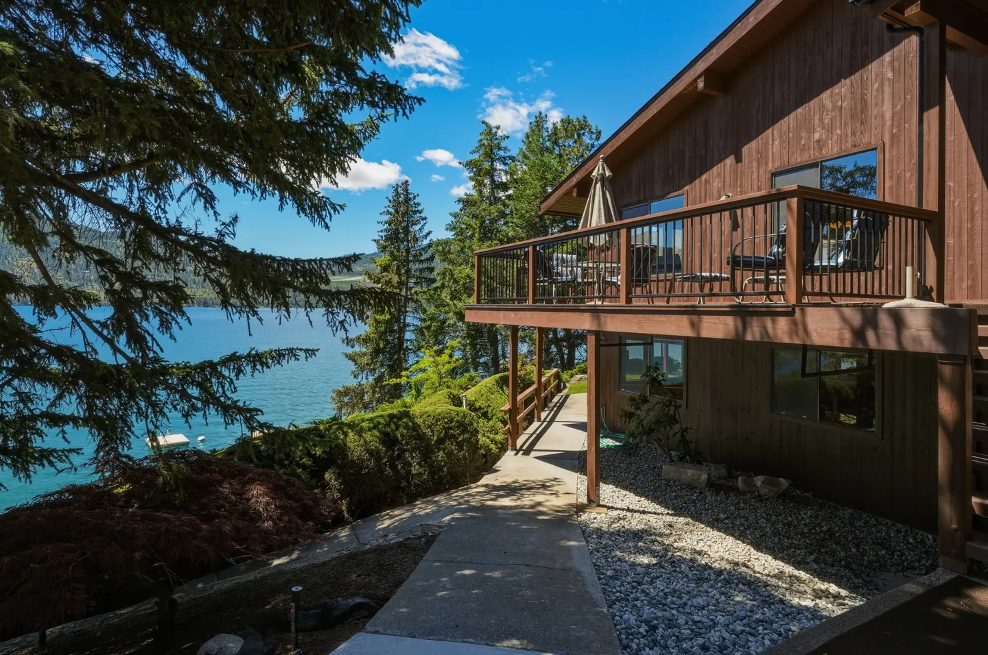 Exterior view of a multi-story wood-sided lakefront house with an elevated deck overlooking blue water and pine trees under a sunny sky.