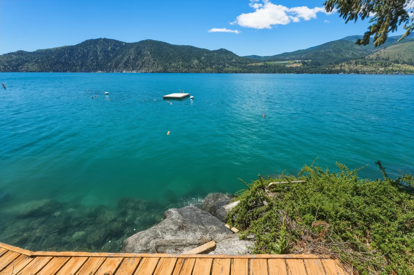 View of vibrant turquoise lake water from a wooden deck, with a floating swim dock and green mountains in the distance.