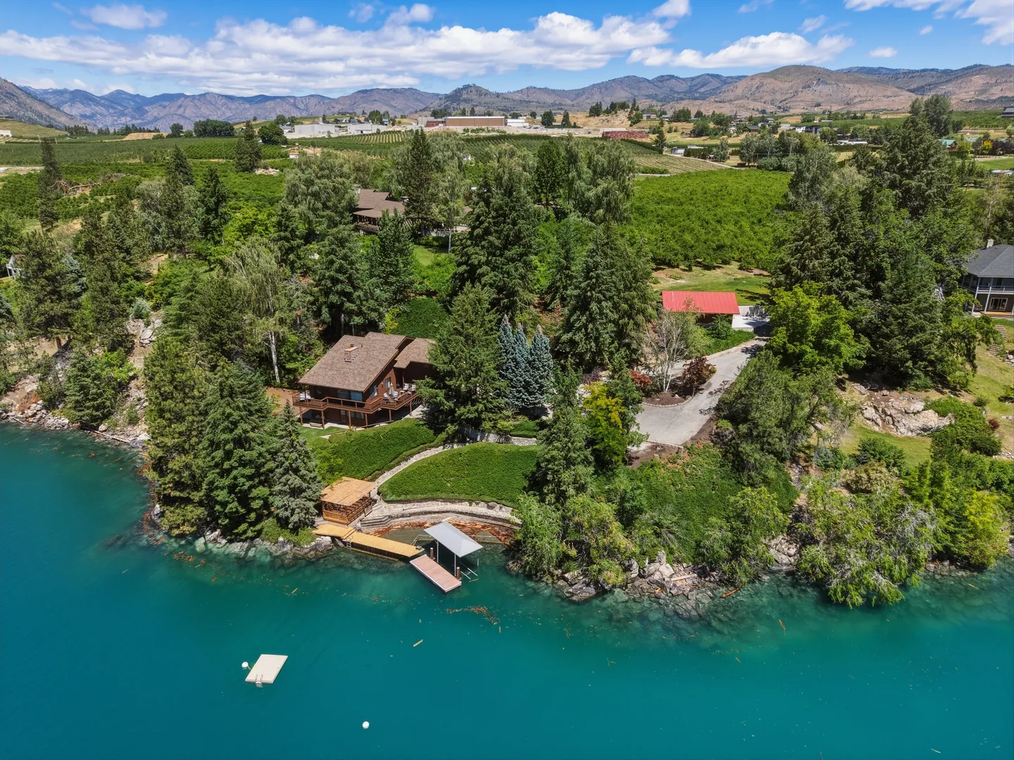 Aerial view of a lakefront house surrounded by pine trees with a private dock, boat lift, and terraced lawn leading to turquoise water.