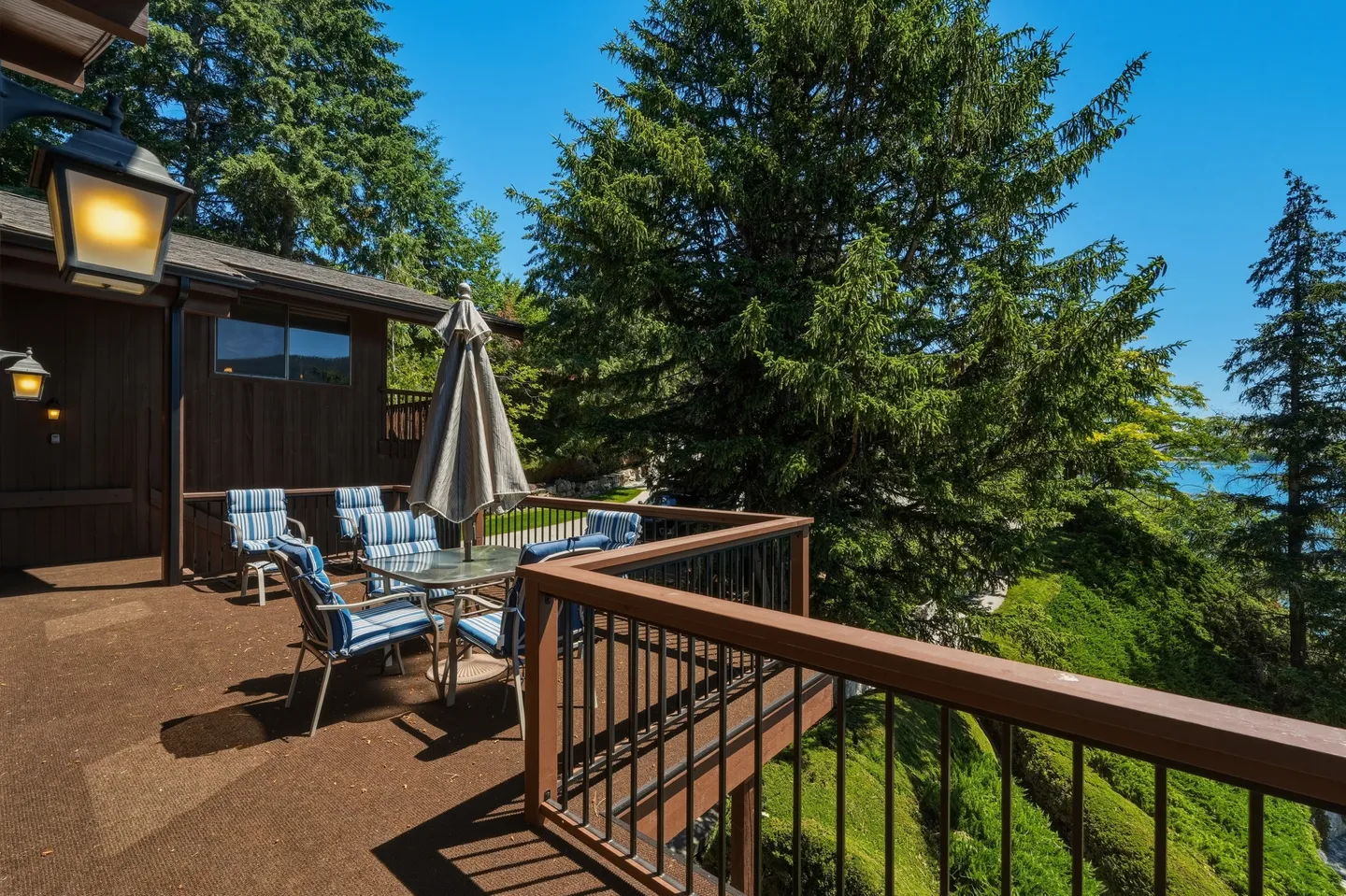Outdoor elevated deck with outdoor dining table, umbrella, and chairs overlooking evergreen trees under a clear blue sky.