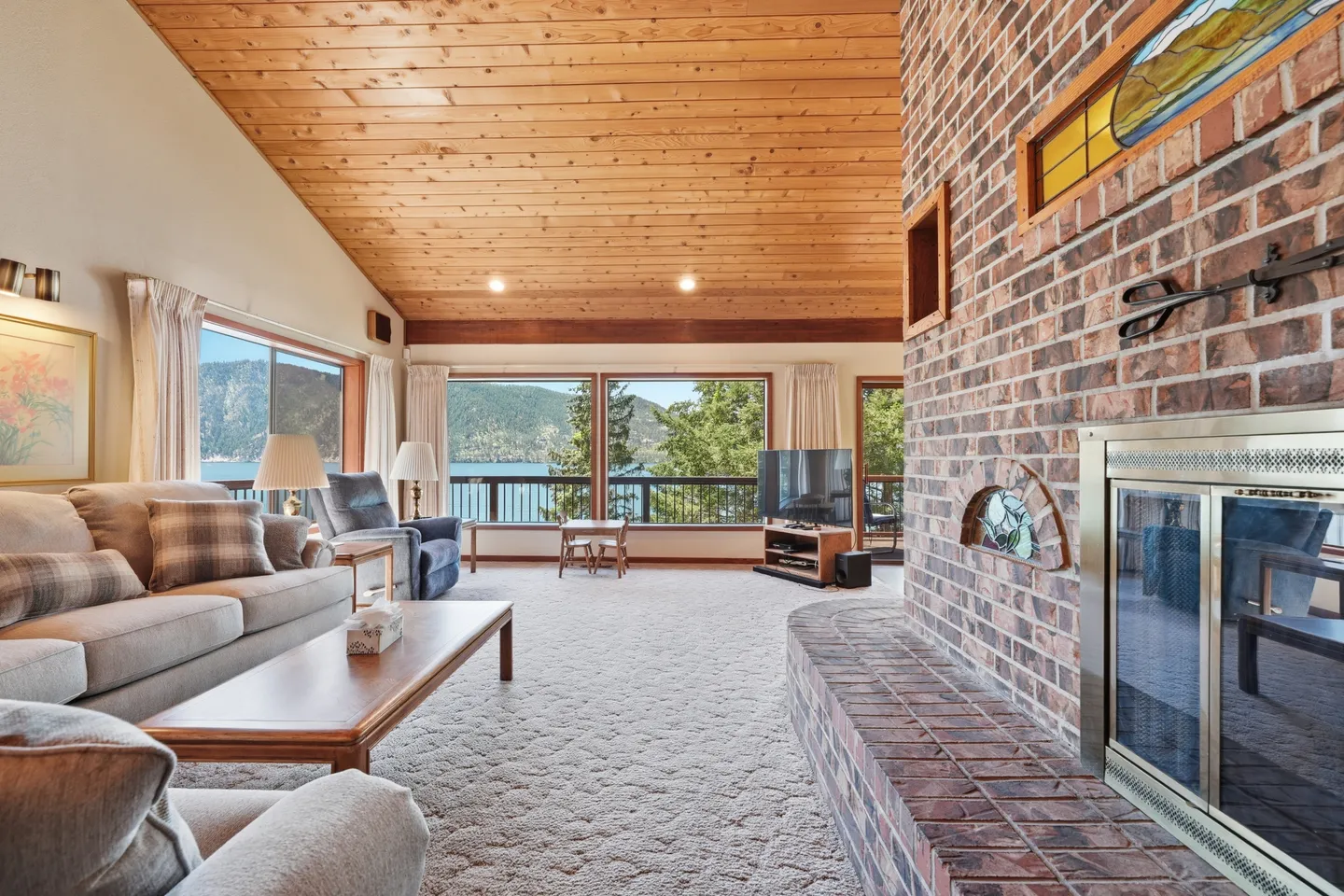 Living room with a large brick fireplace, vaulted wood-paneled ceiling, carpeted floor, sofa seating, and large windows overlooking water and hills.