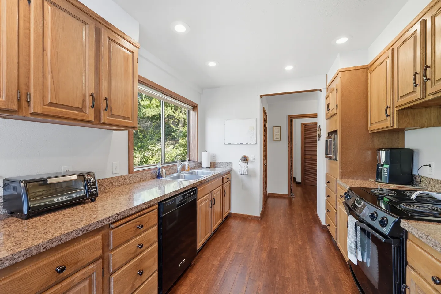 Galley-style kitchen with wooden cabinets, laminate or granite-look countertops, black appliances, wood-style flooring, and a window above the sink.