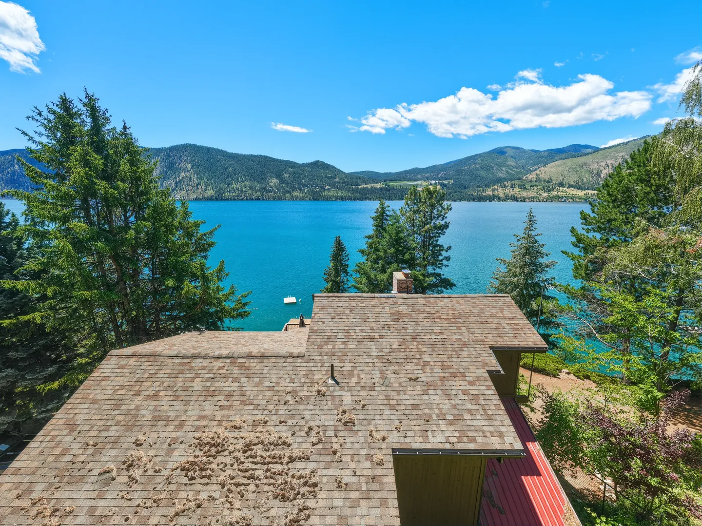 High-angle view over a brown shingled roof looking out onto a bright blue lake surrounded by mountains.