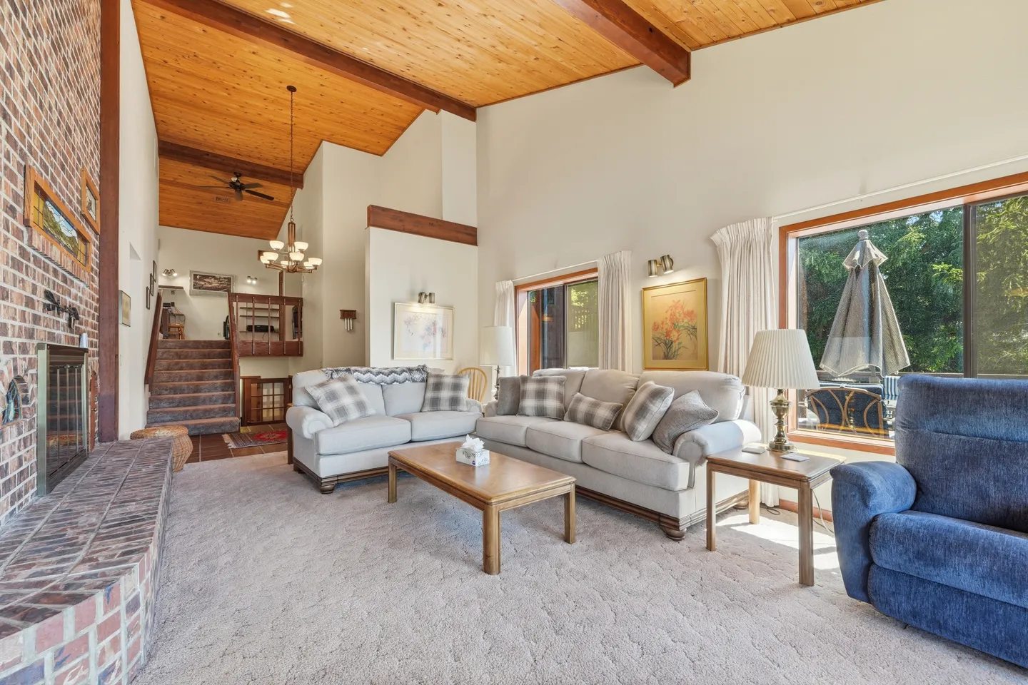 Living room with vaulted wood-plank ceiling, exposed beams, brick fireplace, carpet, and comfortable seating.