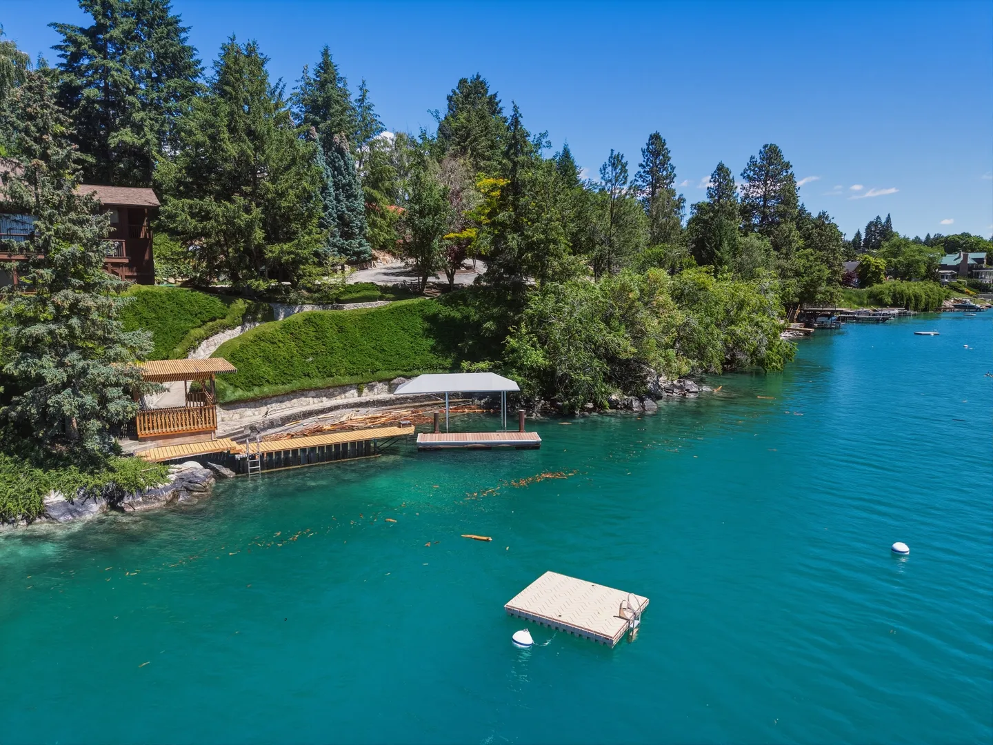 Aerial view of a lakefront shoreline featuring a wooden dock with a covered gazebo, a covered boat lift, and a floating swim platform on clear turquoise water.