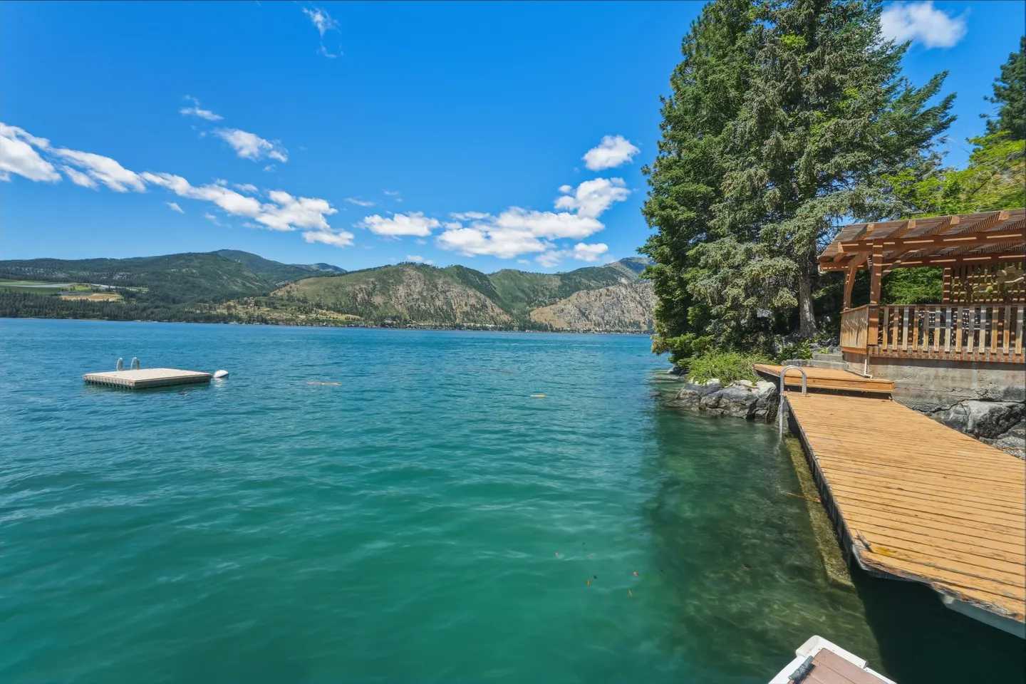 A wooden dock extending into clear turquoise lake water with a covered wooden gazebo on the shore and hills across the lake.