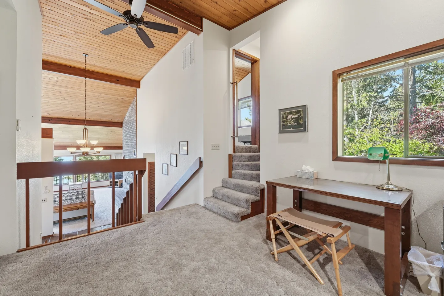 Carpeted loft area overlooking a living room, featuring a small desk with a lamp, wood-paneled ceiling with a fan, and short carpeted stairs.