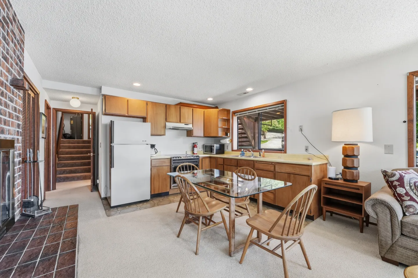 Open living, dining, and kitchen area with carpeted floors, wood cabinets, a glass-top dining table, and a brick fireplace edge.