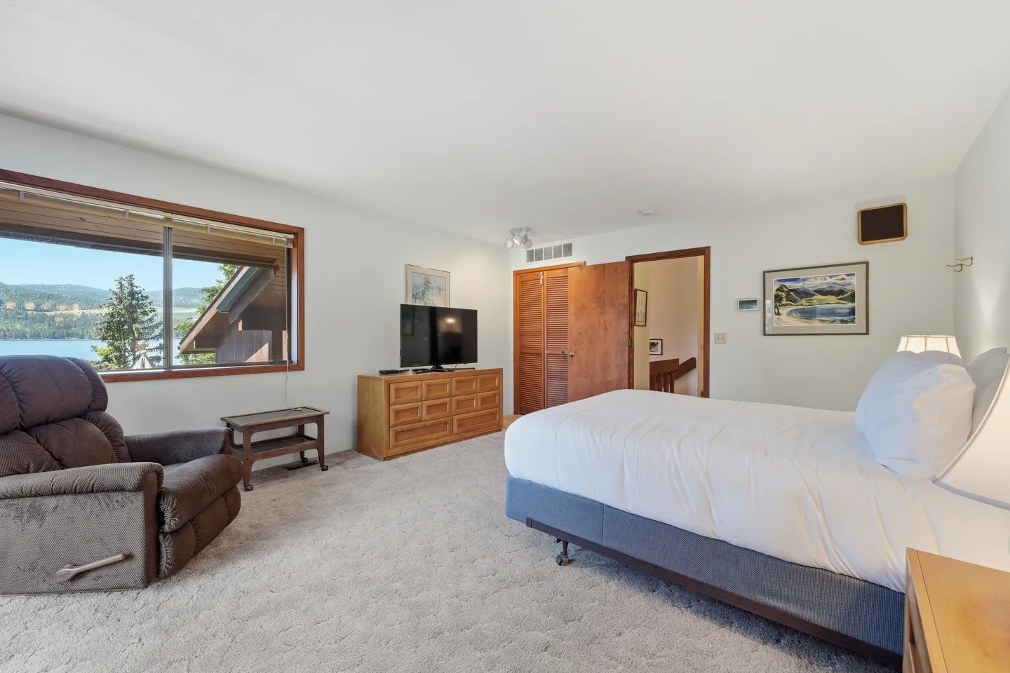 Bedroom with carpet, a large bed, armchair, TV on a dresser, and a window with scenic lake views.