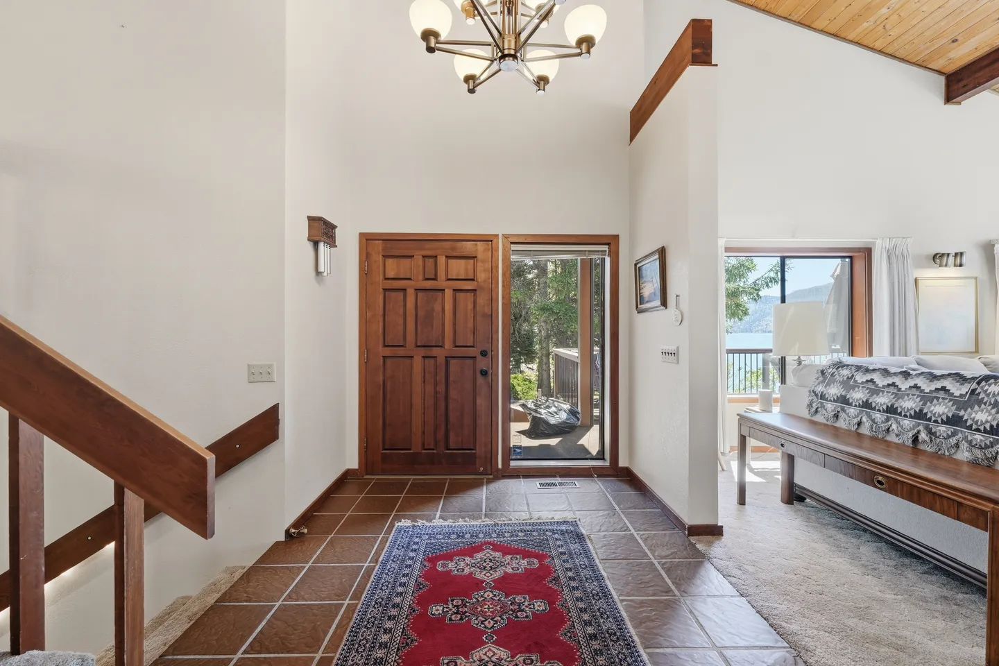Tiled entryway with a wood front door, adjacent glass door, patterned rug, staircase railing, and view into the adjacent living space.