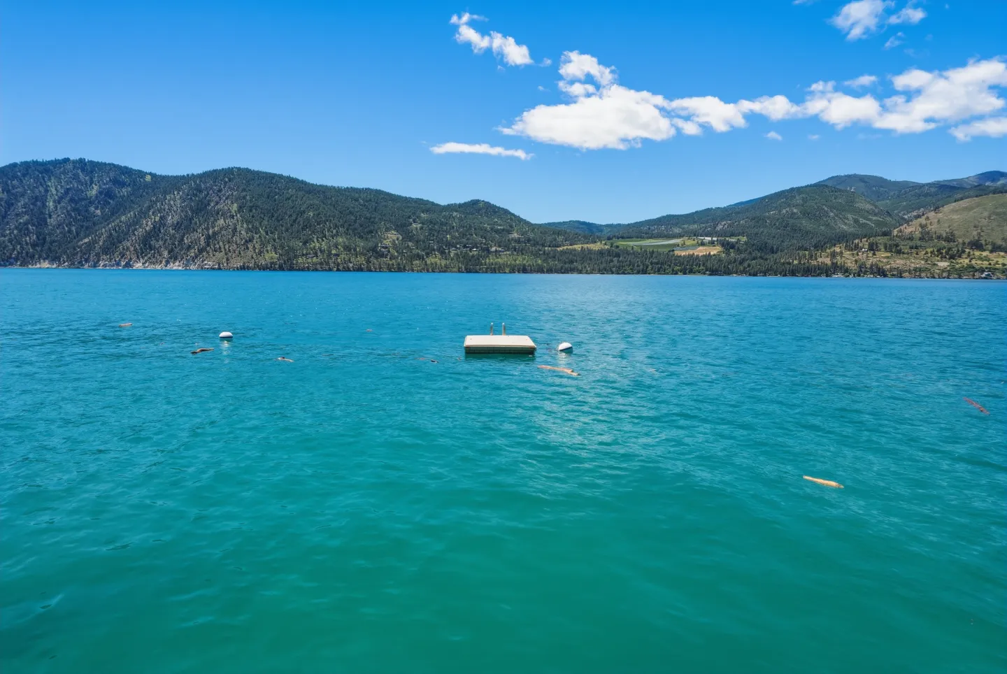 Elevated view of vibrant turquoise lake water featuring a floating swim dock with a swim ladder beneath green hills and a bright blue sky.