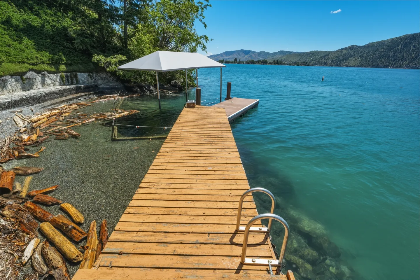 A wooden dock with a swim ladder leading out into clear turquoise lake water next to a covered boat lift under a blue sky.