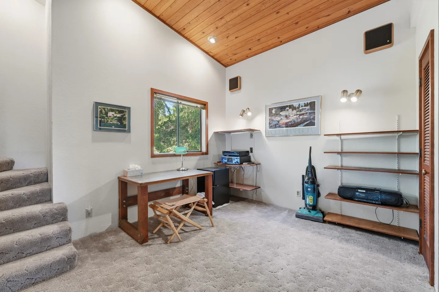 A carpeted room with vaulted wood-paneled ceiling, a desk with a folding stool, shelving, stairs, and a window.