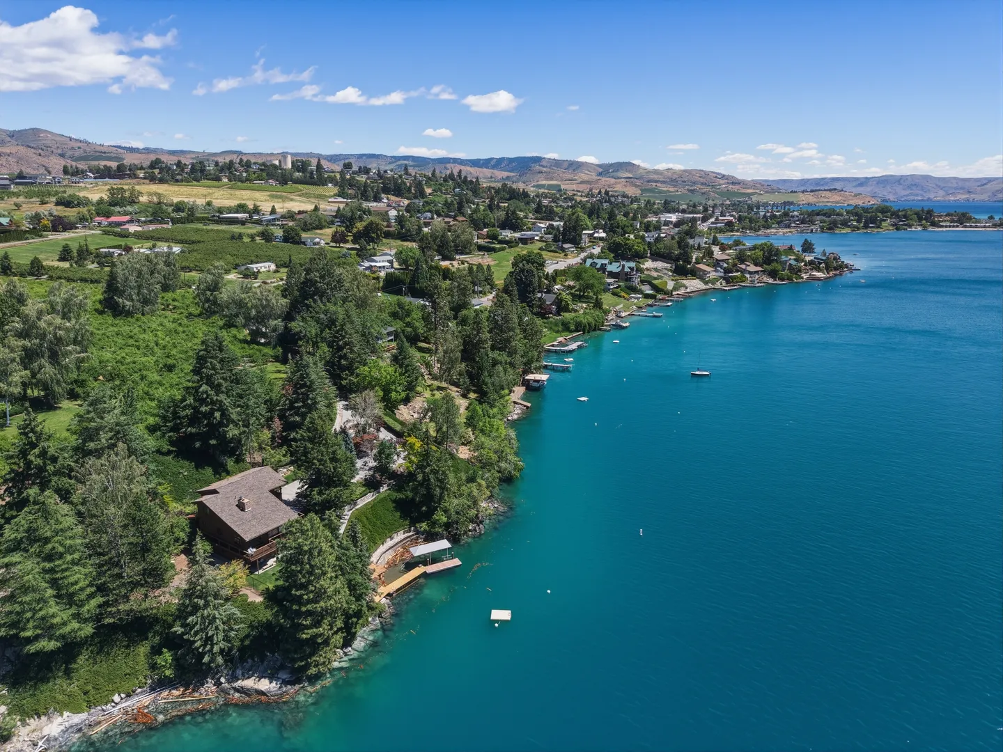 Aerial view of a waterfront home nestled in green trees along a turquoise blue lake with docks and surrounding hillside.