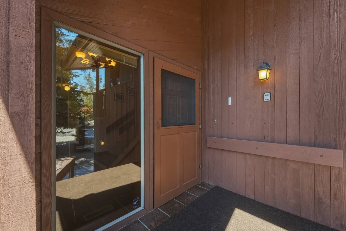 Exterior entryway of a home featuring wood panel siding, a brown front door with a screen, and a large angled window reflecting trees.