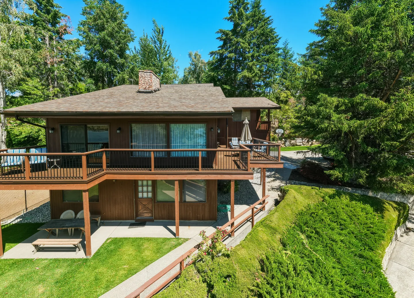 Exterior rear view of a two-story brown wood-sided house with an elevated deck, patio, and lush green lawn surrounded by tall pine trees under a clear blue sky.