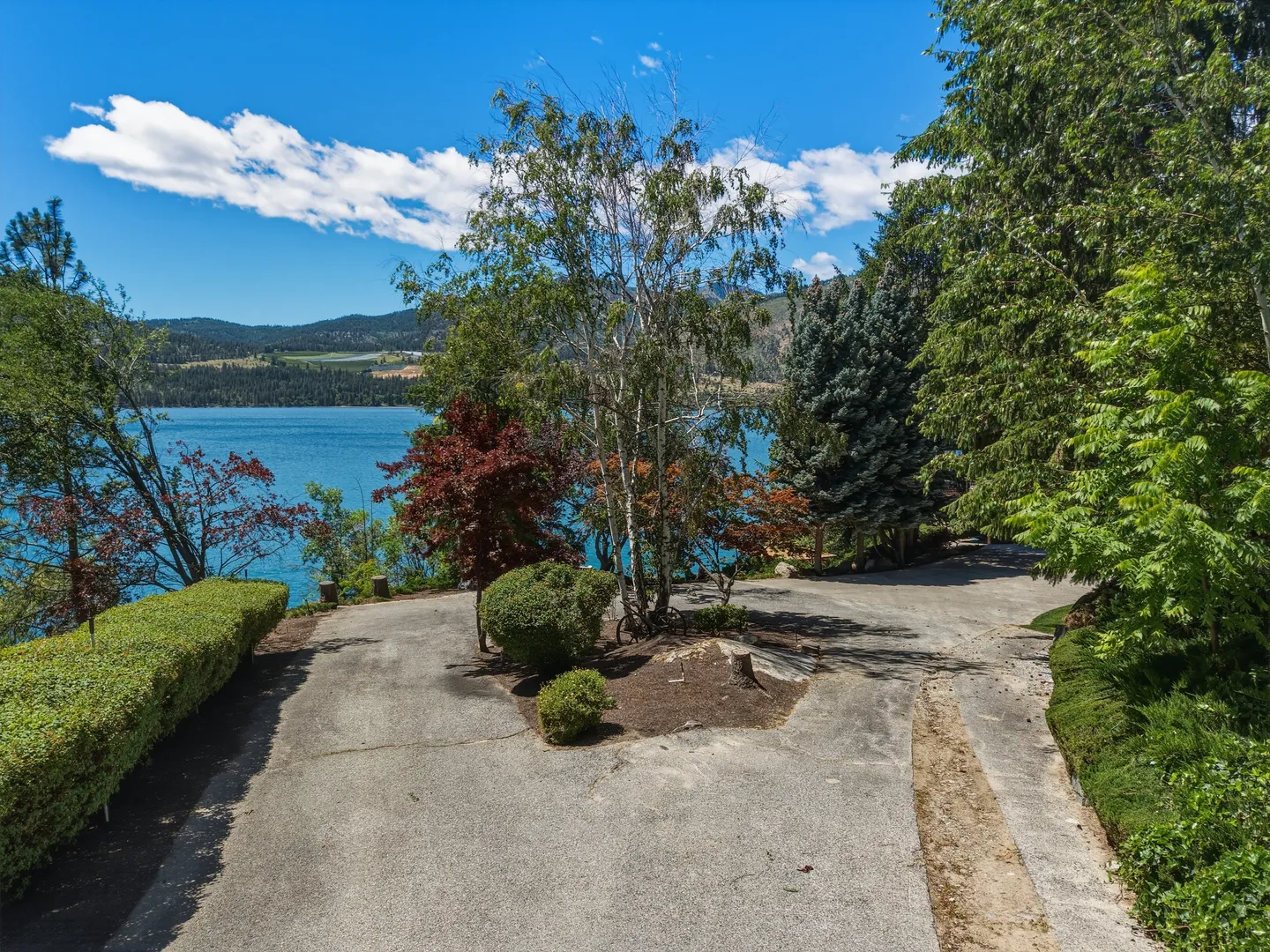 A paved driveway splits around a landscaped island with trees and shrubs overlooking a large blue lake and distant hills under a partly cloudy sky.