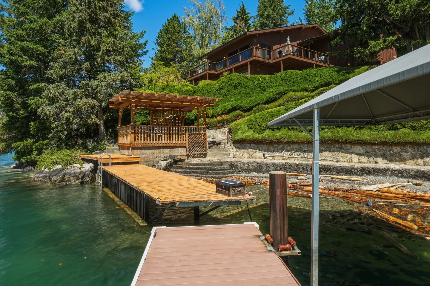 A wooden dock extending into clear water with a gazebo and hillside house visible along the shoreline.