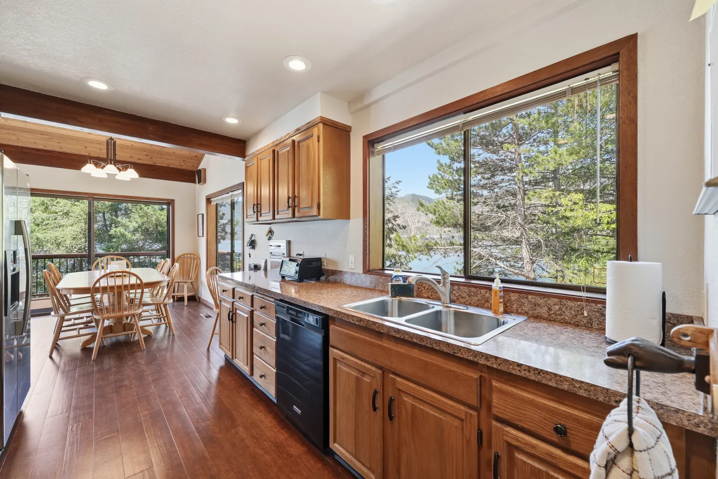Kitchen counter and double sink with a picture window overlooking trees, leading to an adjacent dining area.