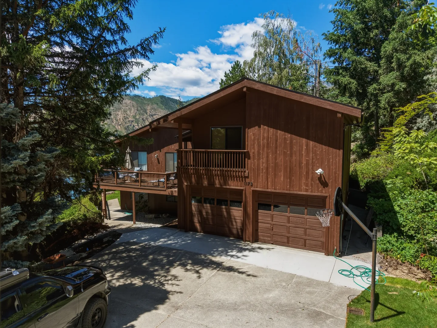 Exterior view of a multi-level brown wood-sided house with a garage, balconies, and surrounding pine trees under a blue sky.
