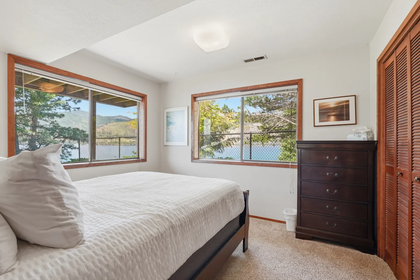 Bedroom with a bed, carpeted floor, dark wood dresser, and two windows offering scenic views of a lake and mountains.
