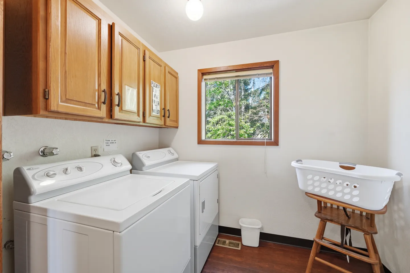 Laundry room with white washer and dryer under wood cabinets, next to a window showing outdoor greenery.
