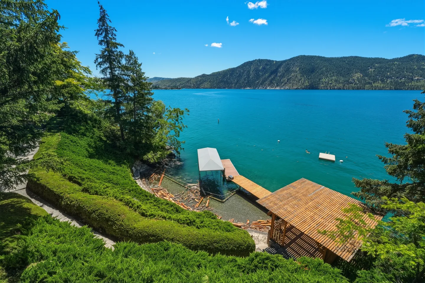 Elevated view of a lakefront shoreline featuring a covered dock, boat lift, wooden pavilion, and lush green landscaping against blue water and mountains.