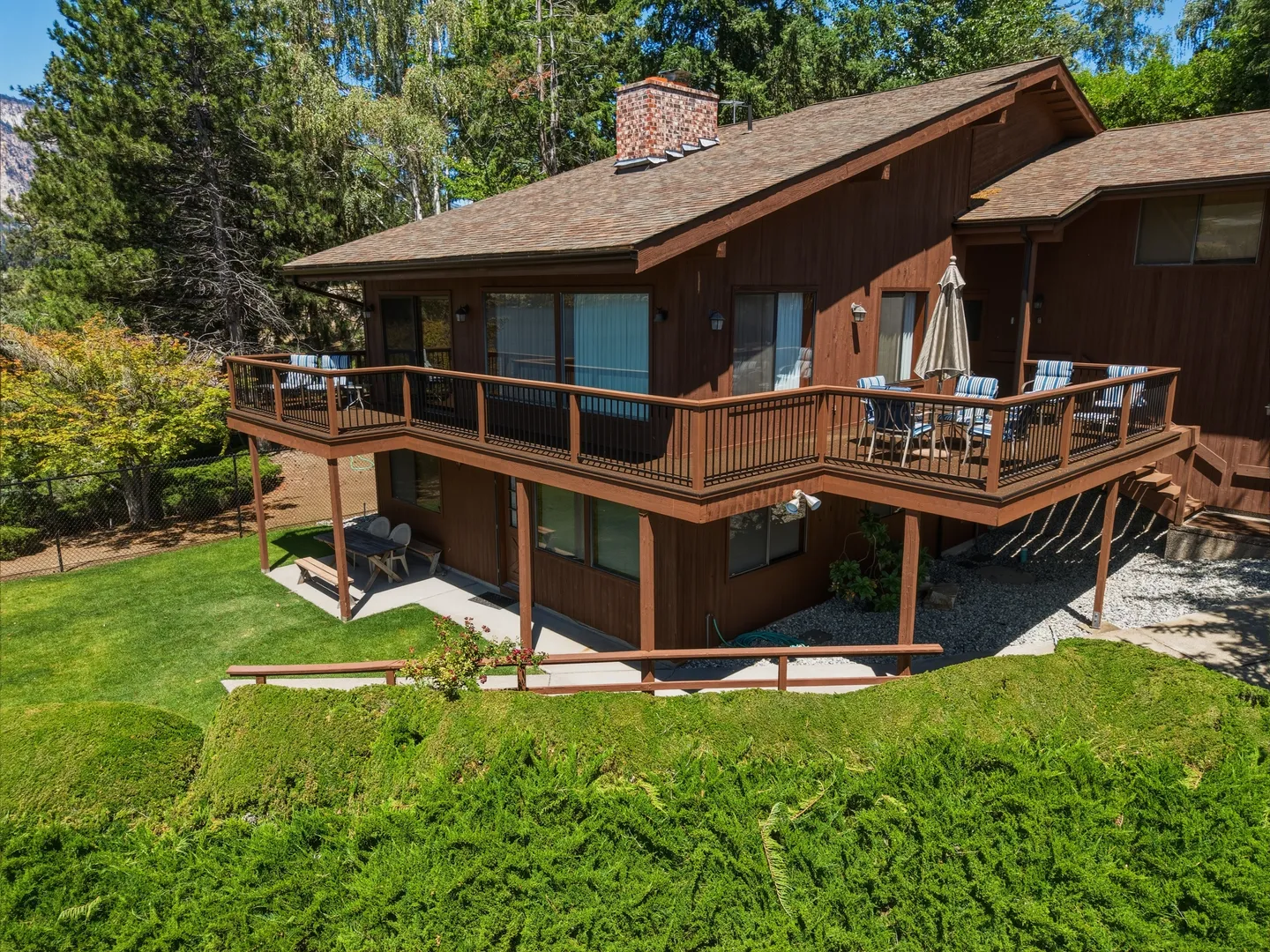 Exterior view of a multi-level wood-sided home featuring a wrap-around upper deck with outdoor seating, a lower patio, and surrounding greenery.