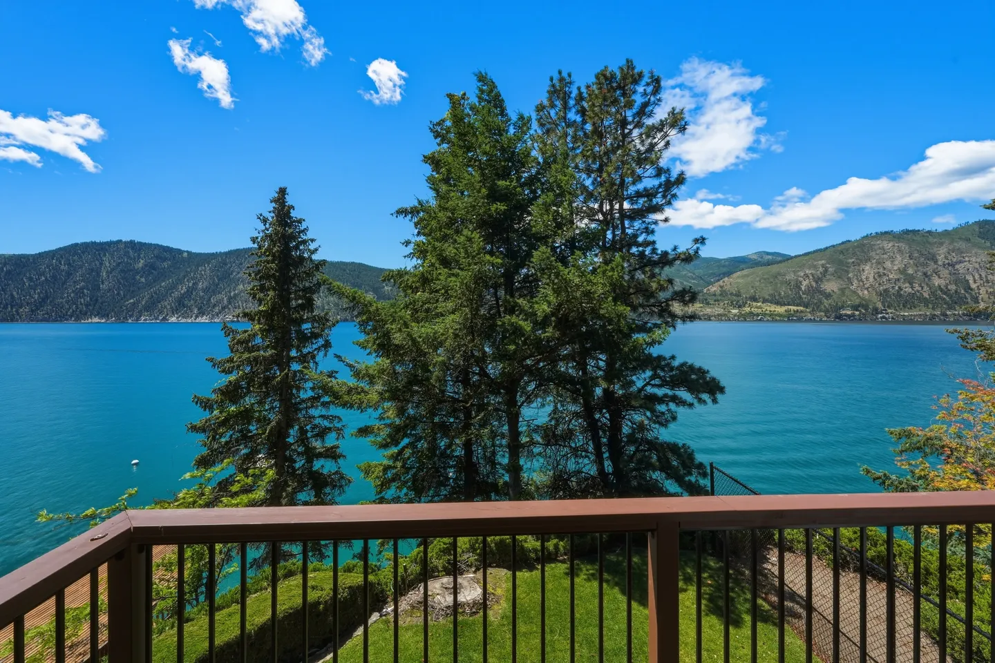 View of a large blue lake and distant mountains framed by evergreen trees from a deck railing.