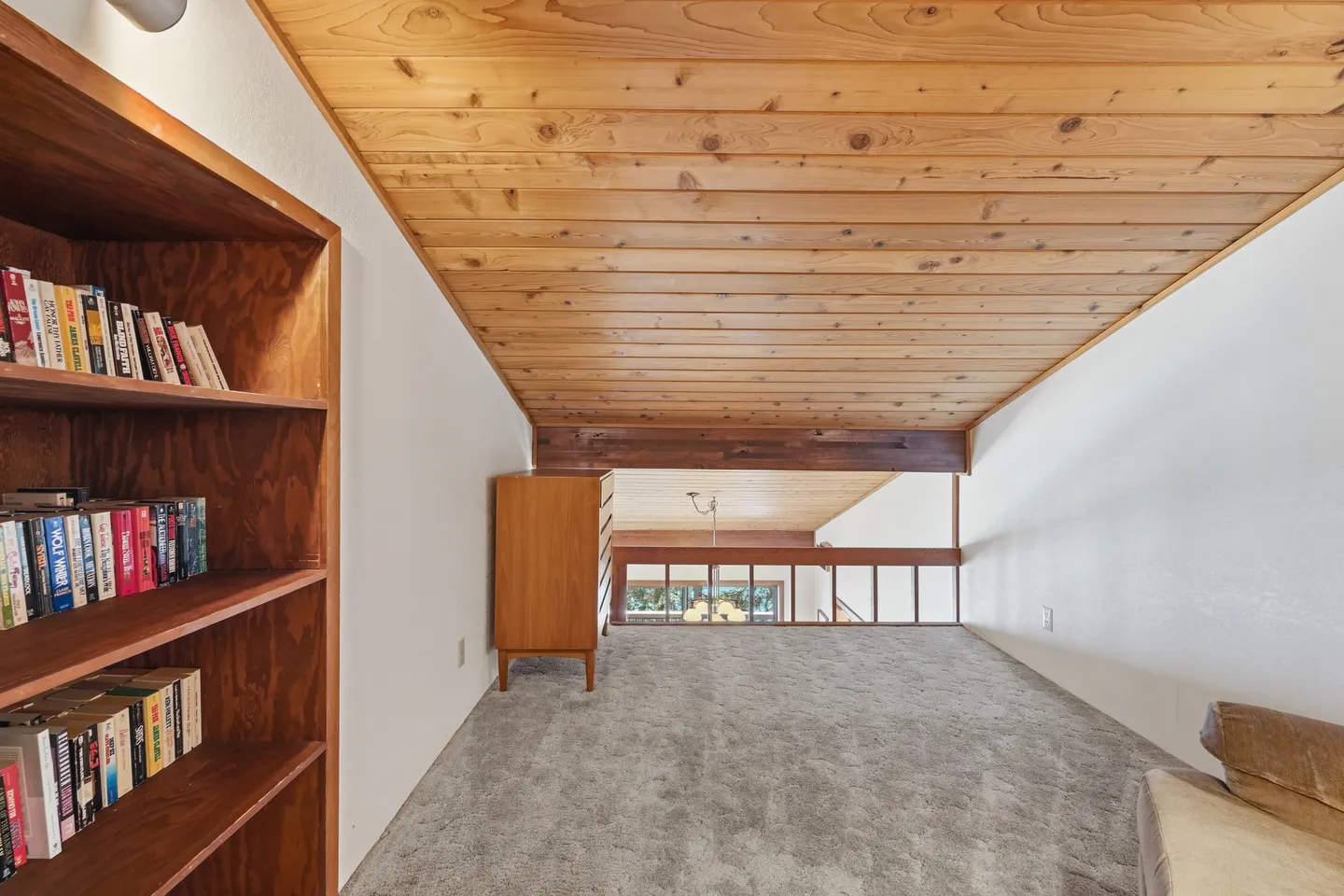 Carpeted loft space with sloped wood-paneled ceiling, bookcase filled with books, and wooden railing overlooking lower level.