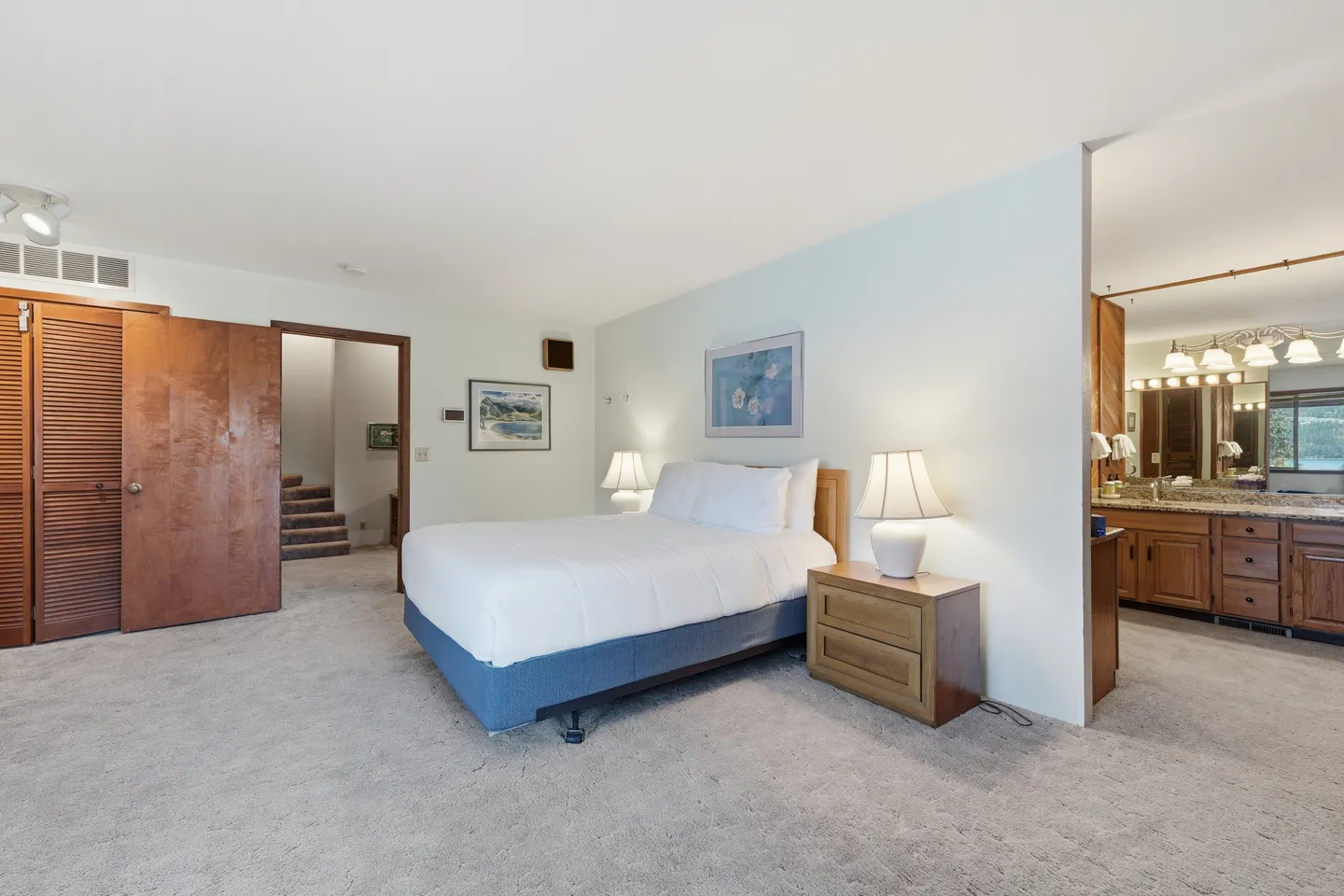 Bedroom with carpet flooring, a bed with a blue base, nightstand with a lamp, and views into an ensuite bathroom and hallway.