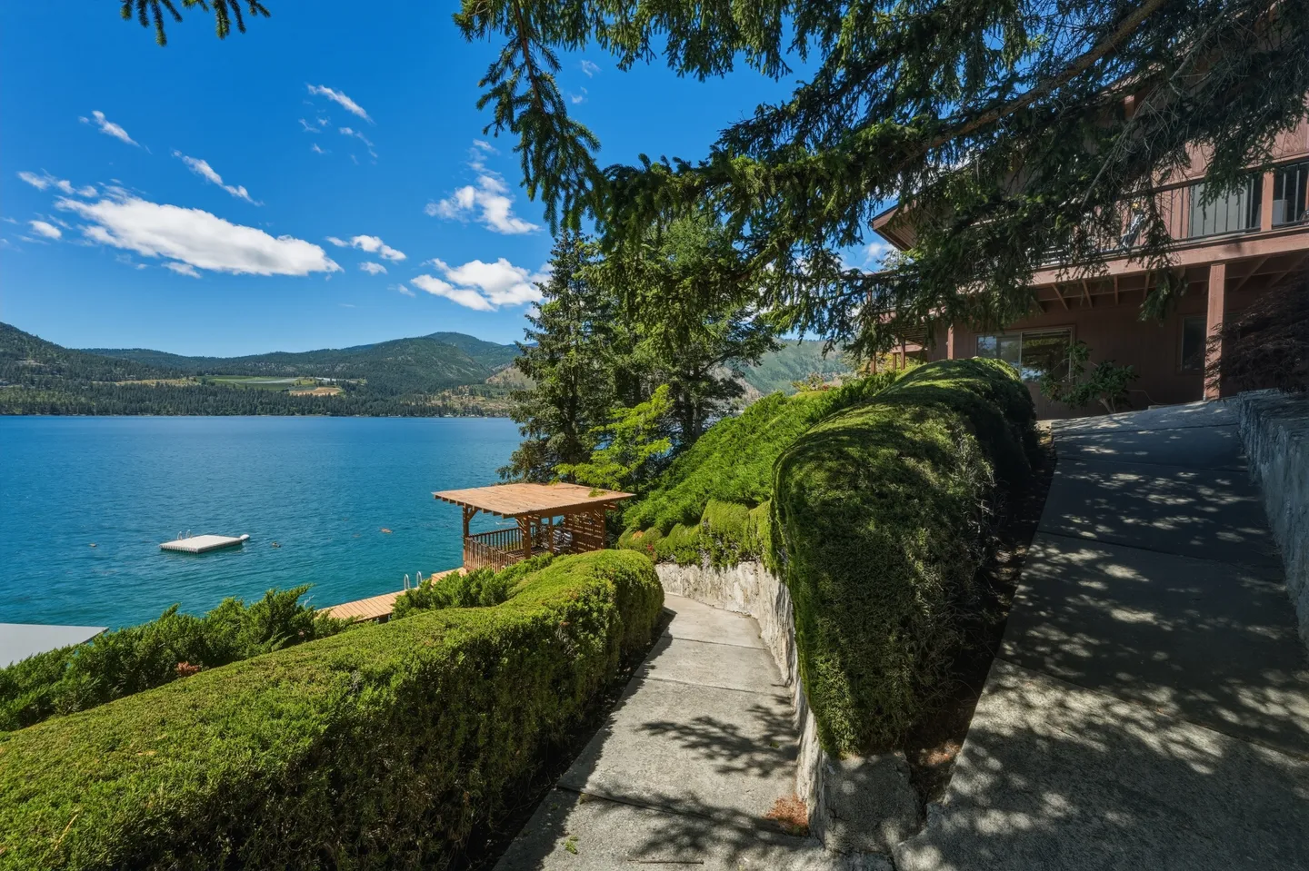 A concrete path lined with manicured evergreen hedges leading downhill toward a lake and dock beside a house.