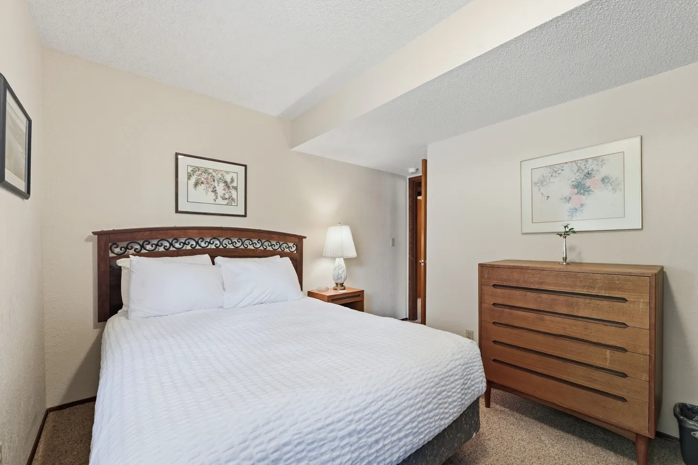 Bedroom with a wooden headboard, white bedding, nightstand with lamp, and a wooden dresser.
