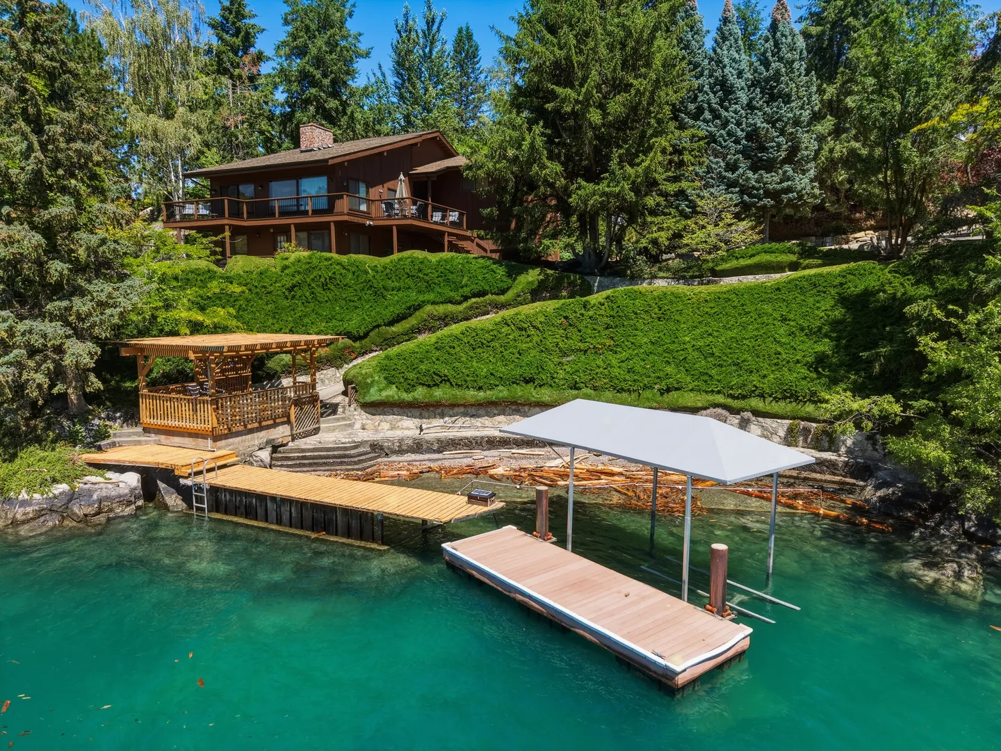 Waterfront view of a multi-story wood home perched on a landscaped hillside above turquoise water with a boat dock and gazebo.