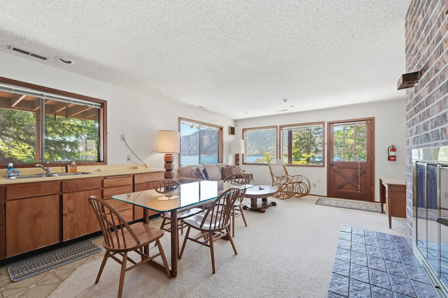 Open living area with a kitchenette counter, dining table, fireplace hearth, and windows overlooking a lake.