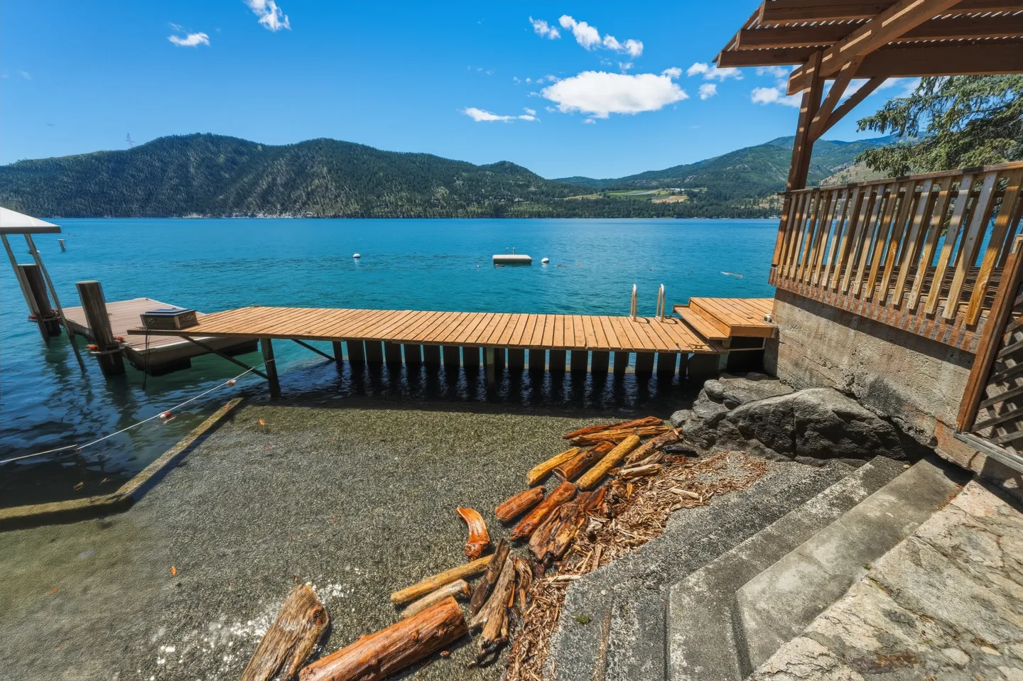 A wooden dock extending into clear blue lake water from a rocky shoreline, with mountains visible in the background under a blue sky.