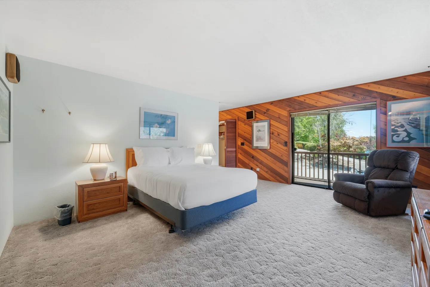 Bedroom with carpet flooring, a bed with bedside lamps, an armchair, and sliding glass doors leading to a balcony.