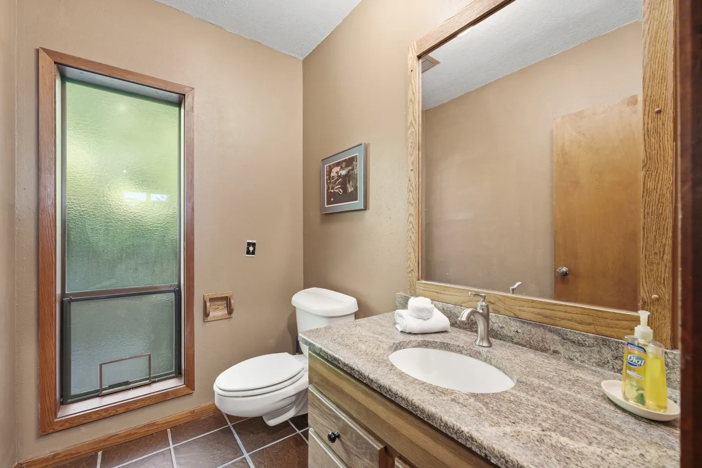 Bathroom with granite vanity countertop, wood-framed mirror, toilet, and frosted glass window with wooden trim.