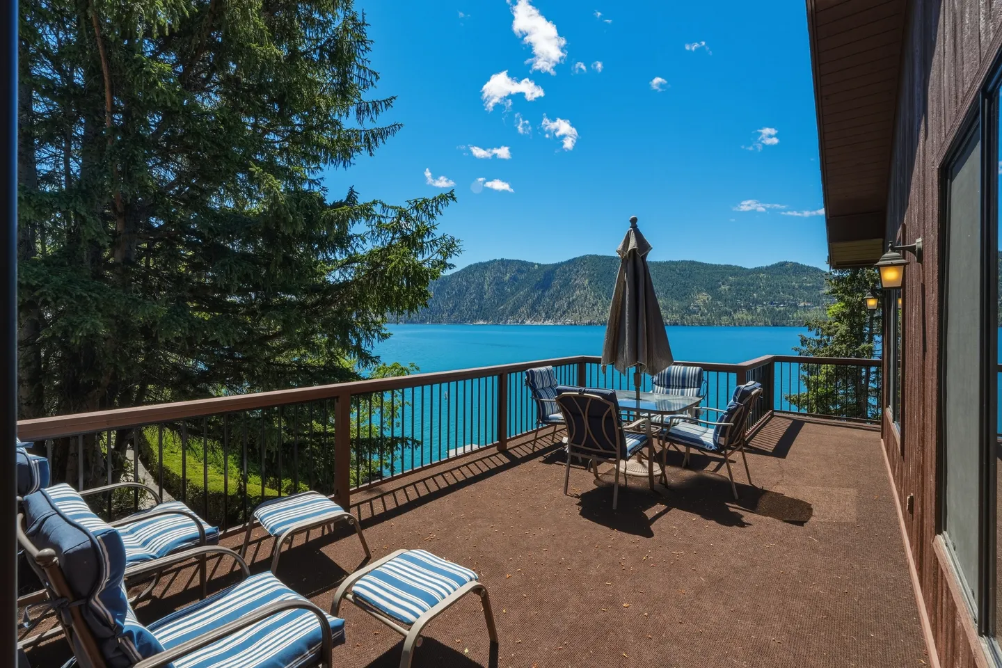 Spacious outdoor deck with patio furniture overlooking a large blue lake and forested mountains under a sunny sky.