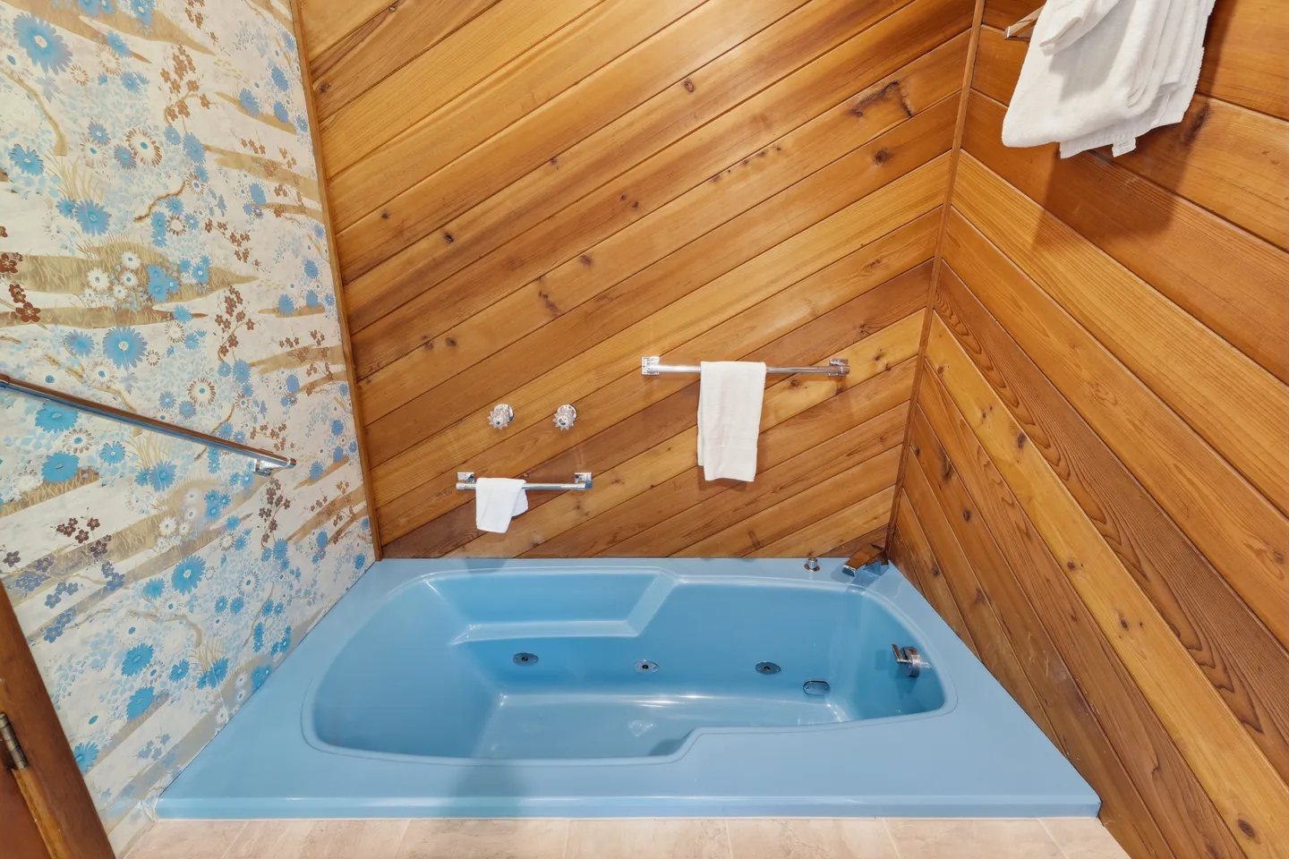 A built-in light blue jetted bathtub in a bathroom with diagonal wood-paneled walls and floral wallpaper.