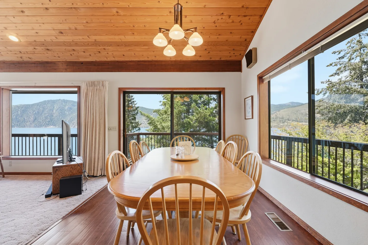 Dining area with wood paneled ceiling, oval dining table and chairs, and windows overlooking a lake and mountains.
