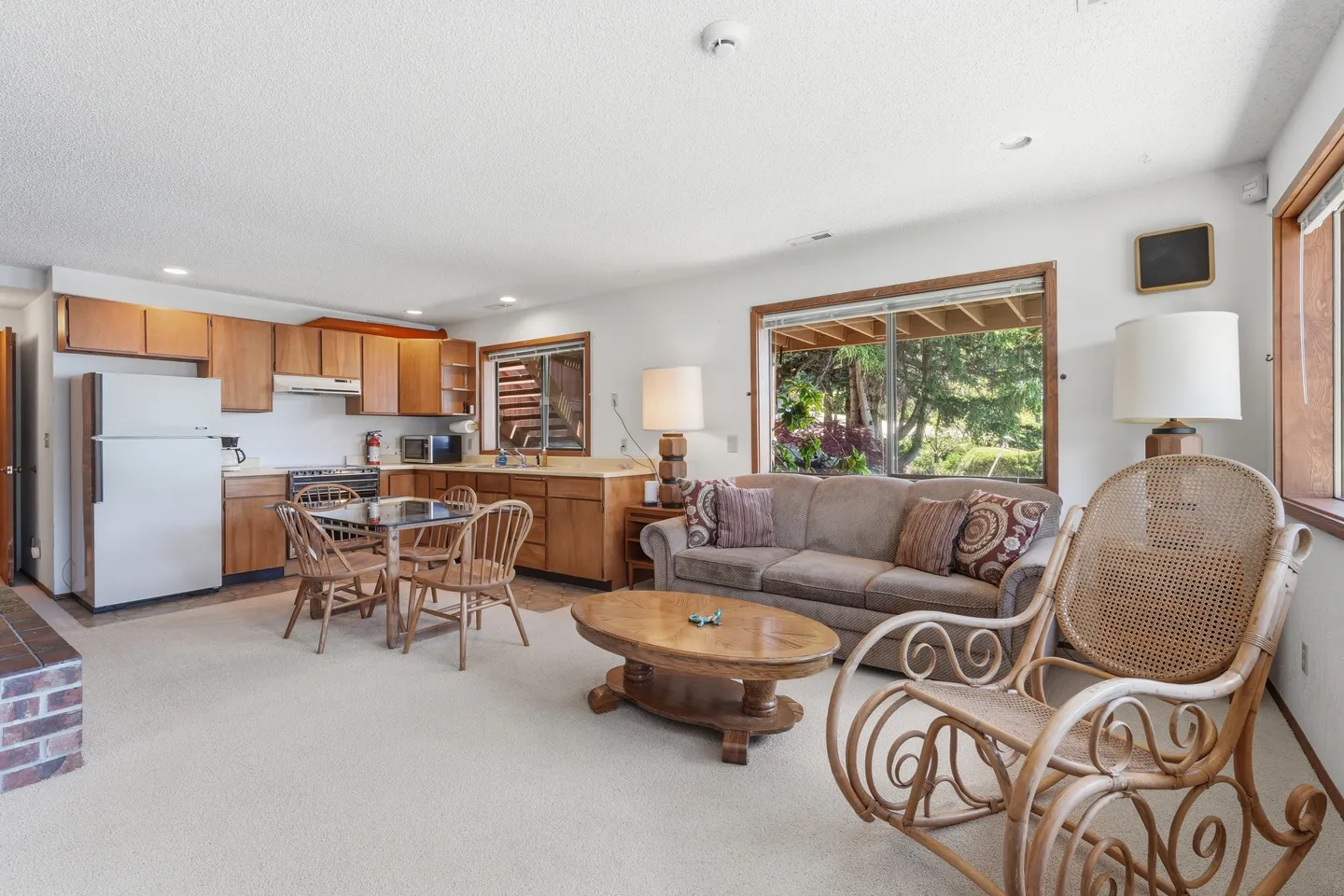 Open living area with carpet, a sofa, coffee table, rocking chair, dining table with chairs, and an adjoining kitchen area with wood cabinets.