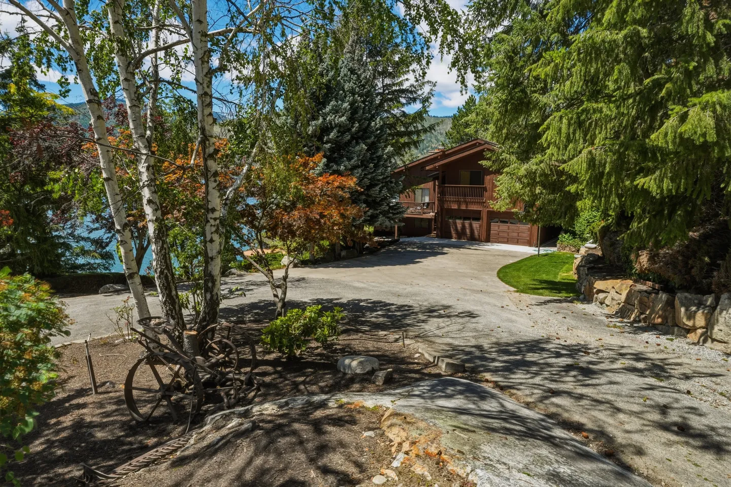 A wide paved driveway leading to a multi-story wood-sided house surrounded by mature evergreen and birch trees next to a lake.