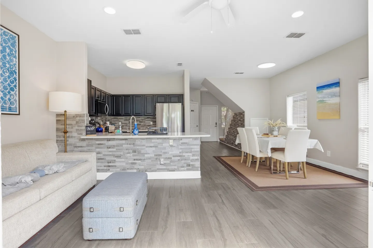 Open-concept living, dining, and kitchen area with grey wood-look floors, a sofa, kitchen island with stone facade, and dining table.