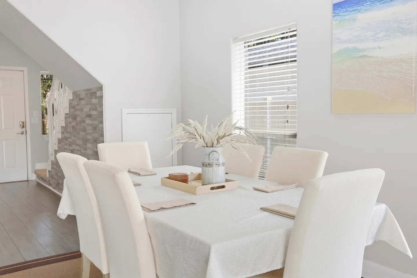Dining table draped in a white cloth with upholstered white chairs, set beside a window with horizontal blinds.