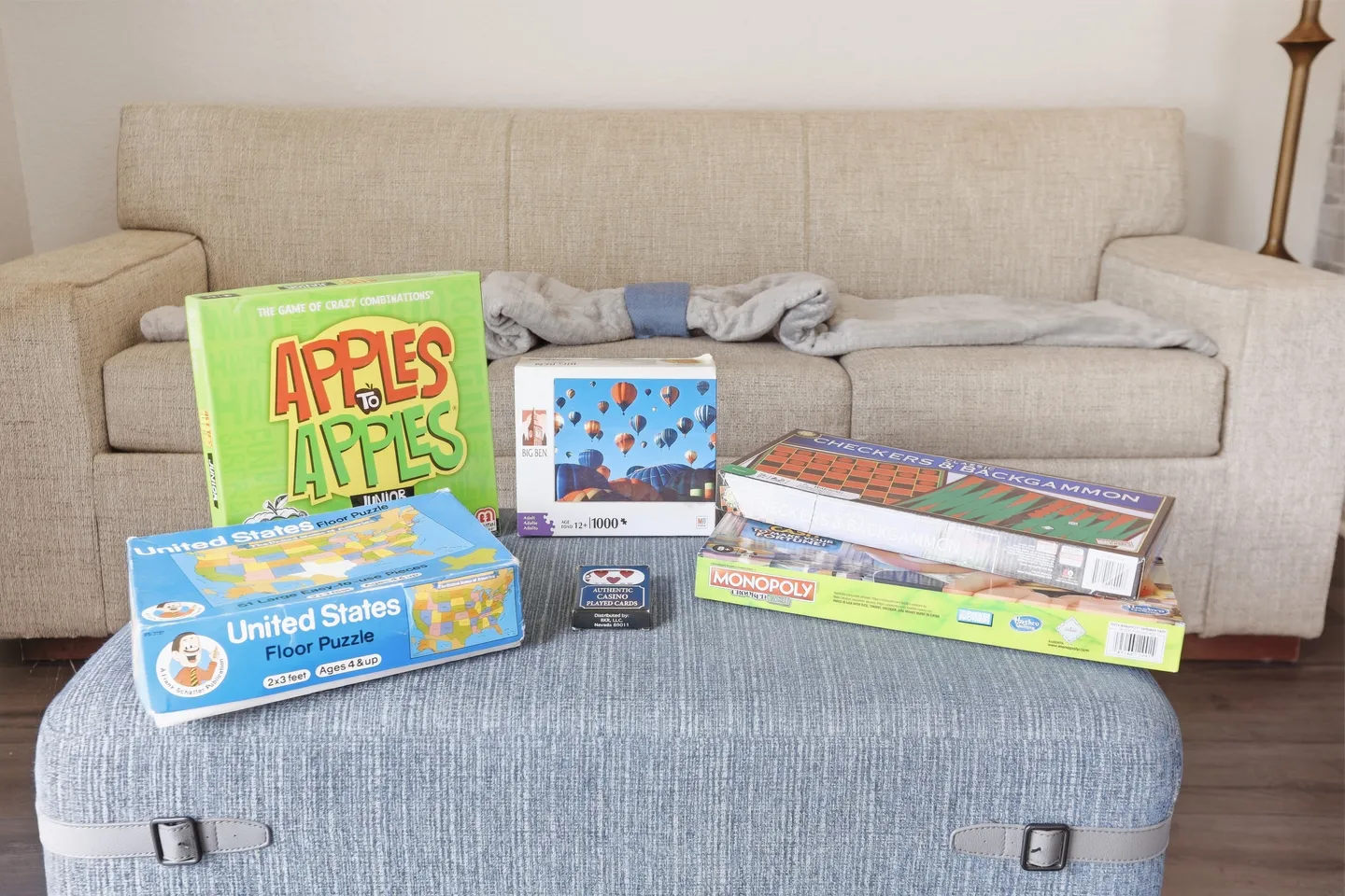 Board games, a deck of cards, and puzzles arranged on a blue fabric ottoman in front of a neutral sofa.