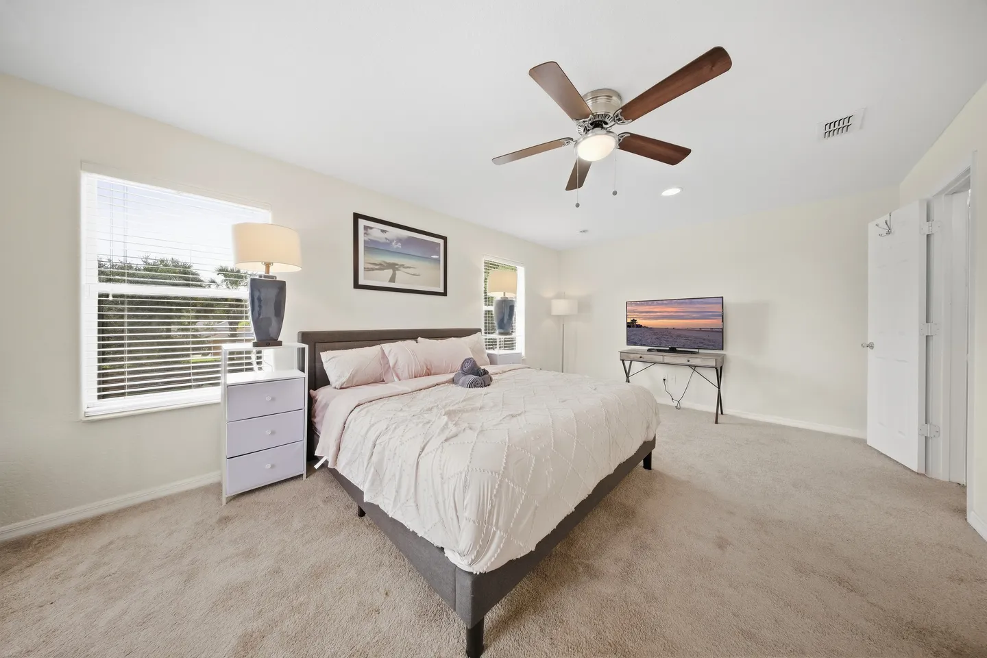 Spacious bedroom with carpeted flooring, a made bed, windows with blinds, a ceiling fan, and a television.