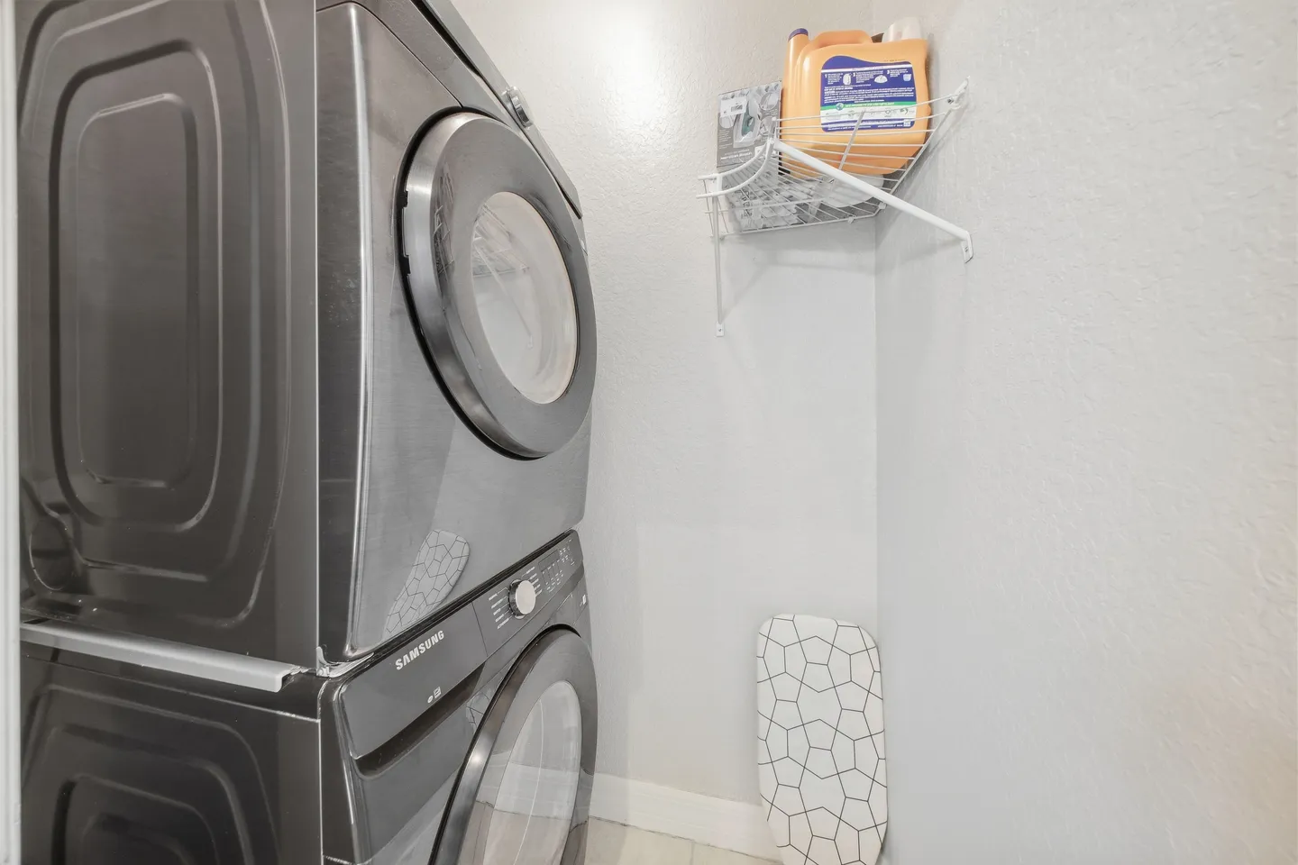 Stacked front-loading washer and dryer in a laundry closet with a wire wall shelf and ironing board.