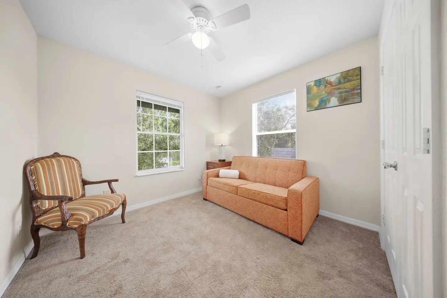A carpeted room with an orange sofa, an antique wooden accent chair, two windows, and a ceiling fan.